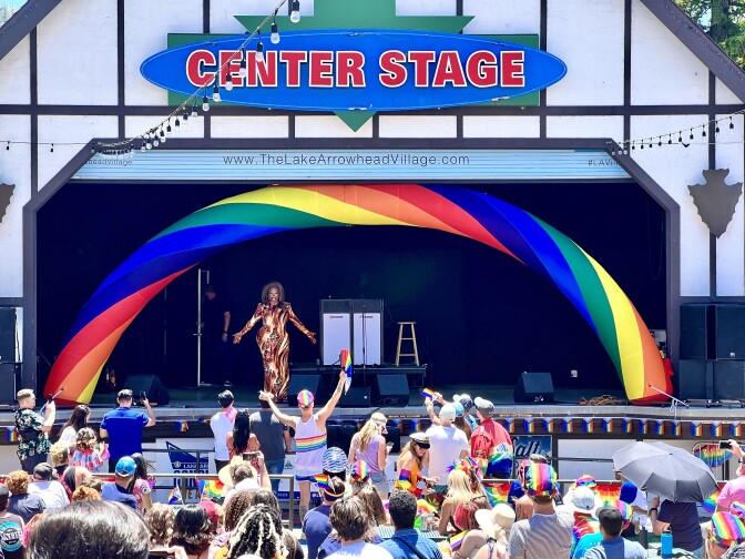 A stake with a rainbow-colored arch over it. Dozens of people in the audience hold rainbow flags and wear bright colors. A drag queen with a long red, yellow and white dress and medium-dark skin tone performs on stage. 