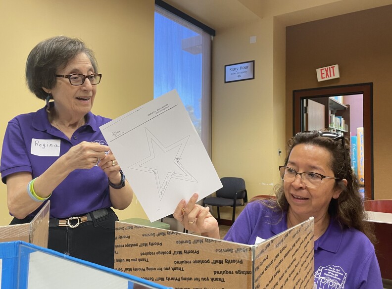 Two women stand behind dividers, both wearing reading glasses. One is holding up a sheet of paper with two stars and some scribbles.