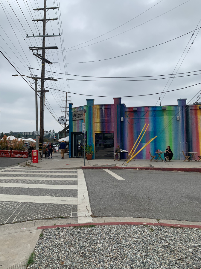 A corner business building is painted in vertical stripes of purple, green, yellow, blue, orange and so on. A few people are walking by, and one person is sitting outside at a patio table.