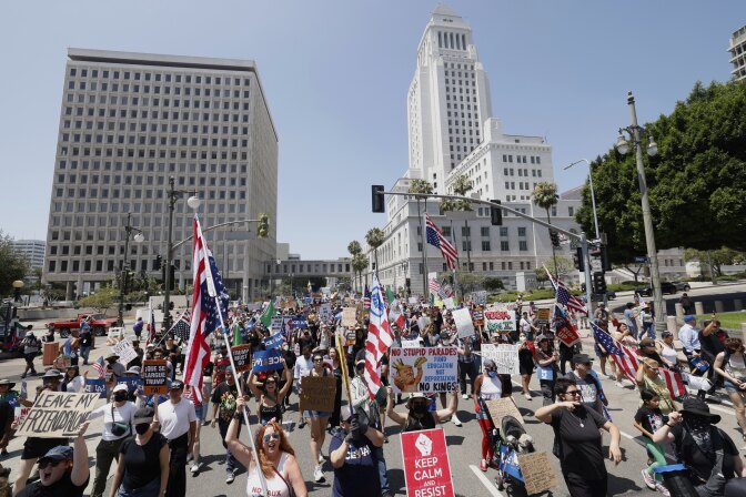 A crowd of people hold signs and American flags as they walk on a street. Buildings are standing behind them with clear skies in the distance.