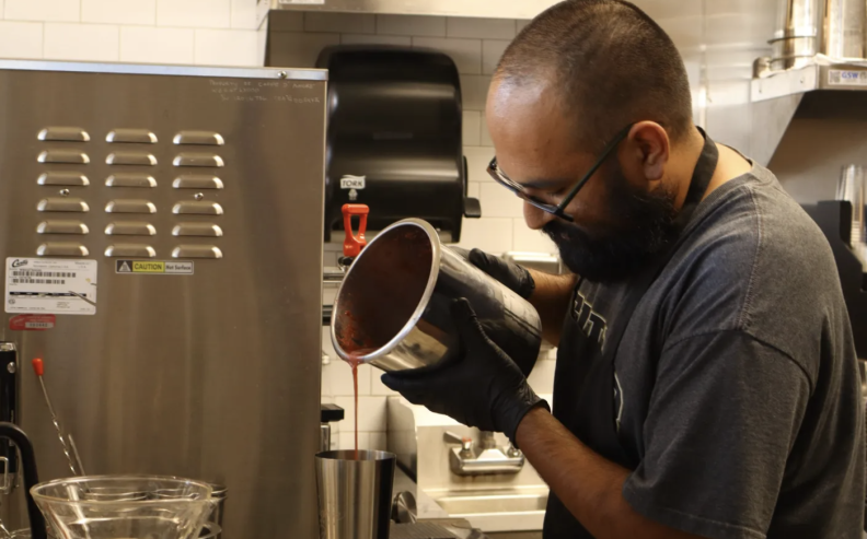 a medium skinner man with a dark beard and glasses, wearing a grey T shirt and a black apron, is concentrating as he pours a bright pink liquid from a large metal canister into a metal cup below