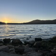 A large blue lake at sunset, with orange and yellow light peaking over a mountain range in the distance. 