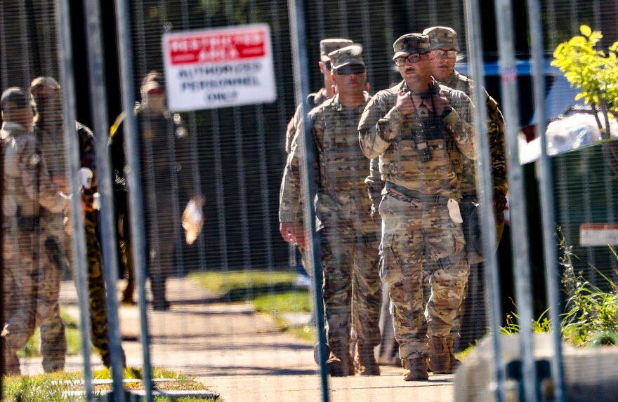 National guard members in uniform walk outside a gated fenced area that is out of focus in the foreground.