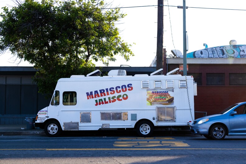 A white taco truck parked on the sidewalk with a red, blue, and green painted sign on its side that reads "Mariscos Jalisco."