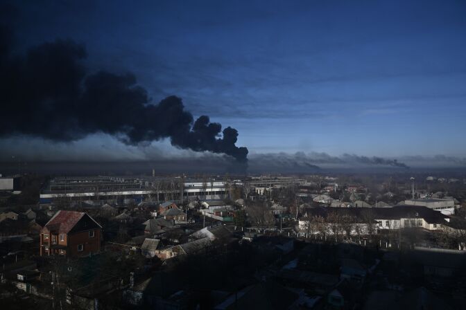 Black smoke rises above a military airport. Low buildings are seen in the lower half of the photo, a large shadow over the lower right portion of the photo. There is smoke rising from other areas further away. The sky remains largely blue with some light clouds, other than the black smoke.