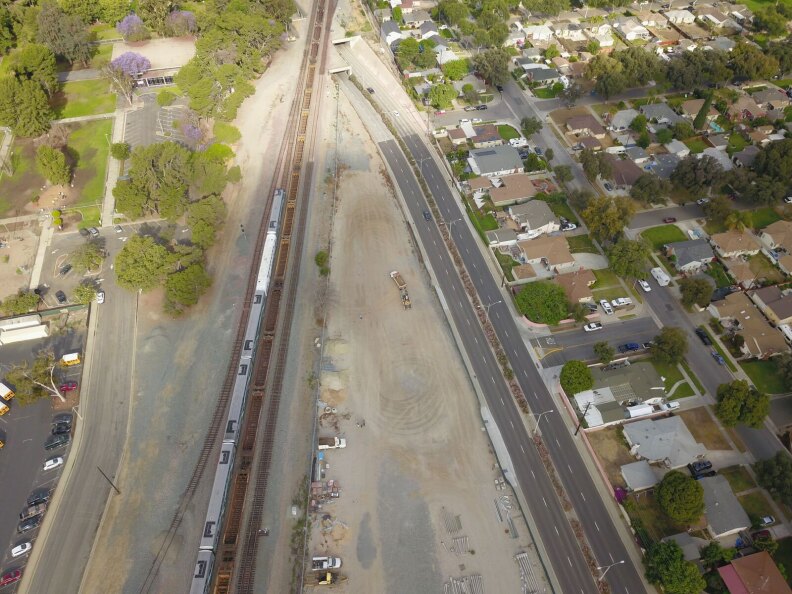 An ariel view of a vacant lot with two paths that come to a point. There is a roadway that goes underground and train tracks that go over it. Next to the roads is a neighborhood.
