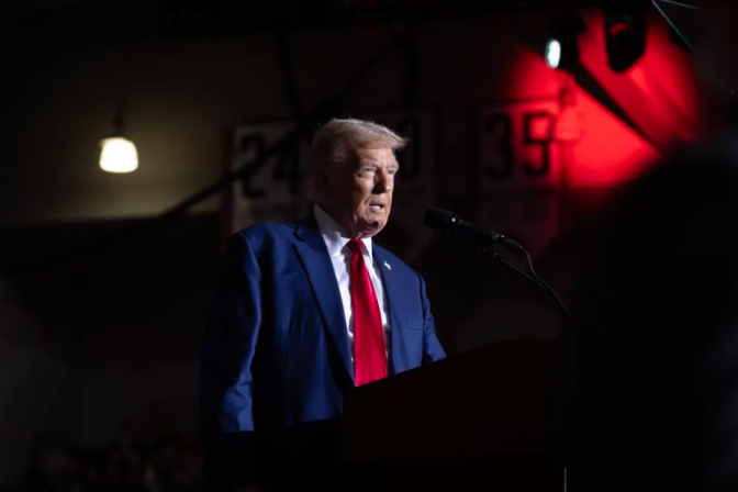 A man wearing a blue suit jacket and a red tie is standing behind a podium.