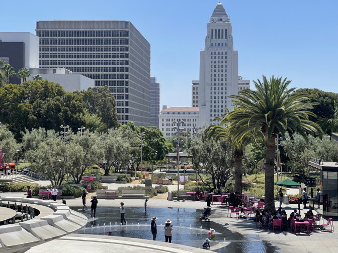 Trees and buildings rise into a blue sky. People stand in a fountain.