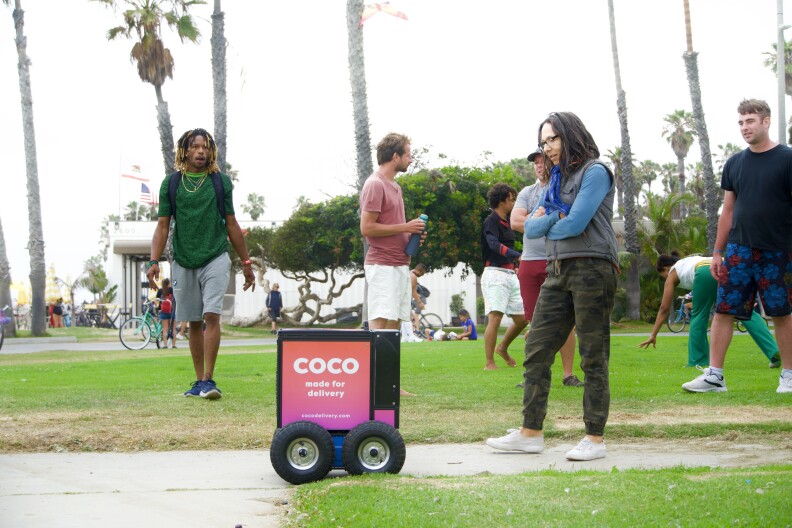 at some sort of park, 3 or 4 people look at a box-shaped robot with wheels while other people hang out in the backgroond
