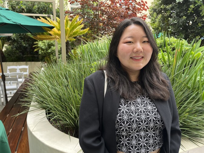 An Asian woman with light skin tone, long dark hair and dressed in black with some white, looks at the camera and smile as she stands in a courtyard, with some green shrubbery behind her.