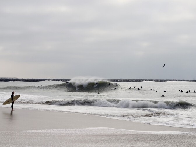A man on shore with his surfboard, in the background are dozens of surfers in black wetsuits, a few of them taking off on a big wave