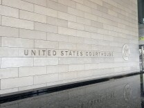 The exterior beige stone exterior has the words "United States Courthouse" engraved with the American federal seal beside it. The letters are reflected from a stone water fountain below the stone exterior wall at the federal courthouse in downtown Los Angeles. 