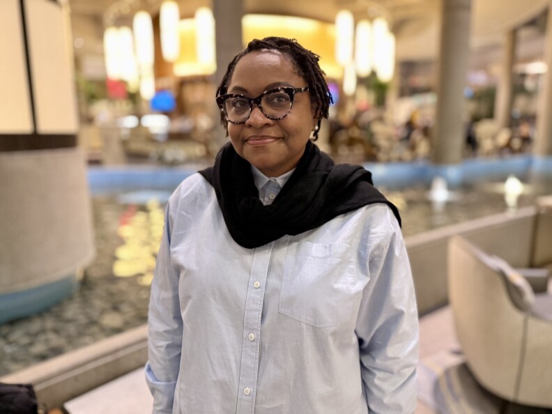 A woman with medium dark skin tone wears a light blue shirt with white buttons and tortoiseshell glasses and has a slight smile.