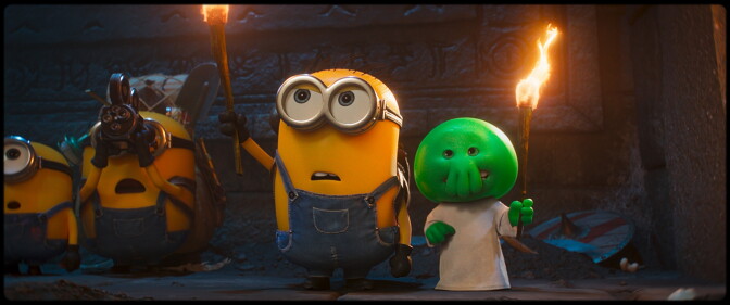 A still of an animated movie, a pill shaped yellow creature wearing overalls holds up a torch, two more creatures like him are in the background. A green smaller creature holding a torch is also present.