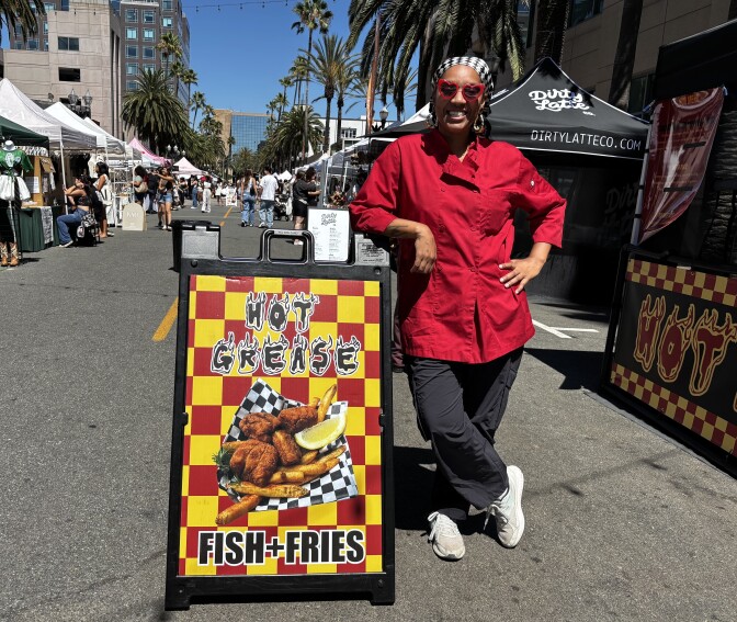 A woman with a dark skin tone wears a bright red chef coat and heart-shaped sunglasses. She stands with her elbow on a stand which says “Hot Grease fish and fries." 