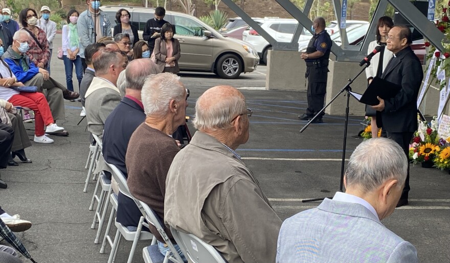 Reverend Albany Lee is dressed in black and standing at a microphone. Behind him are bouquets of flowers. Members of the Laguna Woods and larger Orange County are seated in rows of folding chairs set up in the church parking lot.