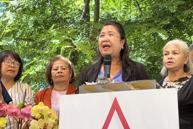 A 65-year-old Thai woman with long dark hair and glasses stands at a podium outside while three other women of Asian descent flank her. 