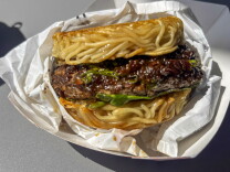 A close-up of a ramen burger — a beef patty glazed in dark sauce with arugula, sandwiched between two seared noodle buns — resting in white food paper.