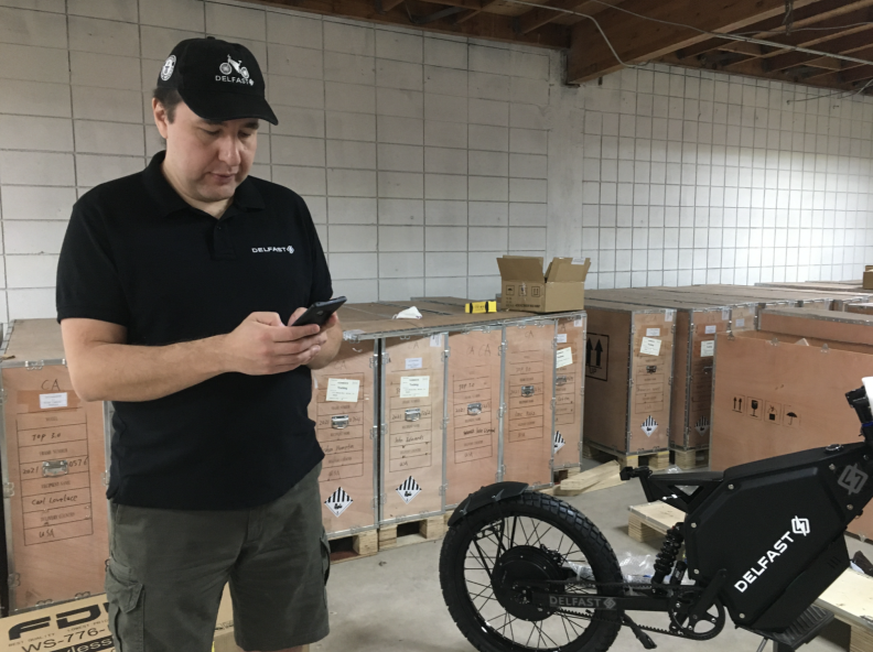 A man in a black polo shirt, surrounded by boxes and a black electronic bicycle, on his cell phone.
