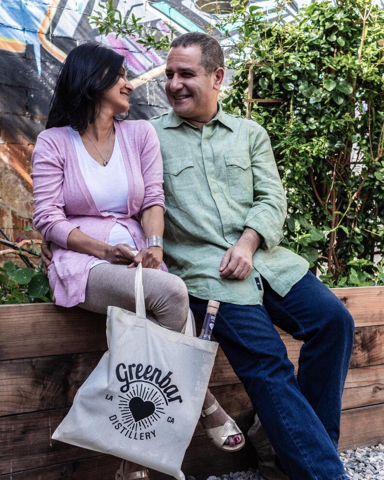 A man and woman sit close on a garden bench, grinning at each other. She has dark skin and black hair, while he has medium-light skin with grey hair. A Greenbar tote with a peek of a bottle rests between them, sunlit and easy, against a backdrop of greenery and street art.
