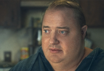 Brendan Fraser sits right of frame, only his shoulders up are visible. Fraser wears a gray shirt, his hair thin and stringy on top of his head. He looks off to the left. His expression is sad, his eyes slightly hooded under faintly furrowed brows. His lips are slightly parted. Behind him seems to be a kitchen with brown cabinets and clutter on the countertops. 