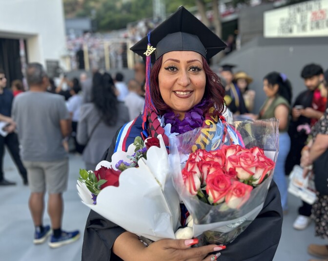 A woman with medium skin tone and long burgundy hair smiles while holding two bouquets of flowers. She is clad in a black cap and gown and drenched in colorful stoles and ribbons. 