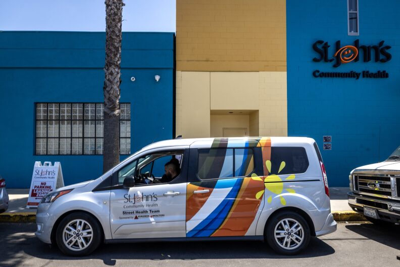 A white mini van with a yellow flower and an orange, blue and white pattern painted on it, is parked in front of a teal colored building