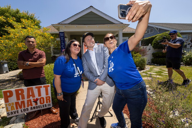Spencer Pratt, a man with light skin tone, wearing a gray suit and black hat, takes a selfie with two woman wearing blue LA Dodgers shirts in front of a home with a lawn sign that reads "Pratt for LA Mayor. Save Los Angeles." Two people stand around them, one recording with a camera.
