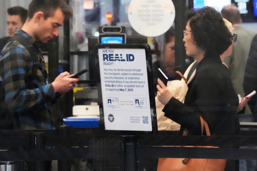 People wait in line in an airport. A small sign on a poll in the line reads "Are you REAL ID ready?"