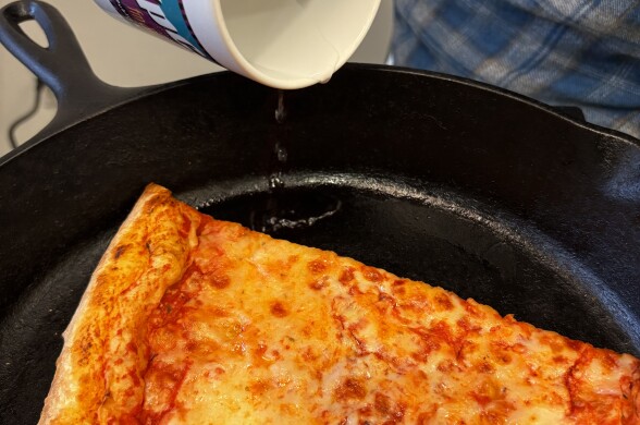 A white mug pours water into a black cast iron pan with a single slice of cheese pizza inside of it