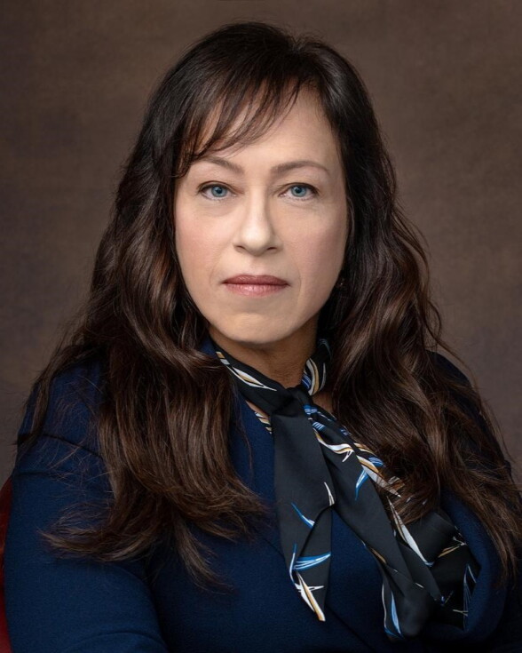 A white woman with dark brown hair sits in a leather chair. She is wearing dark blue, and has a patterned scarf tied around her neck. 