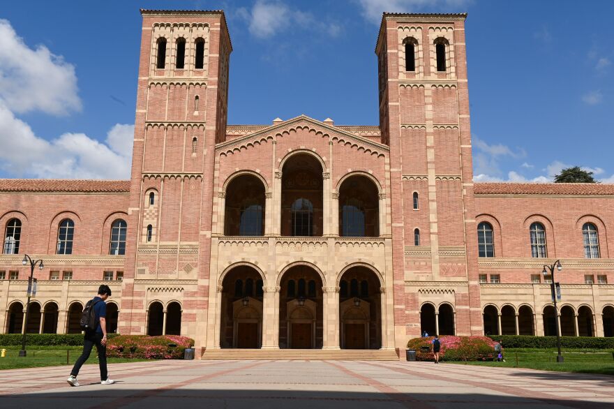 A large brick building at UCLA. A student with a backpack walks nearby, as does another young woman in a dress.