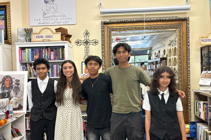 A group of students stand in a bookstore with their arms around one another. In the background, a small gray sign reads, "The Untold Story." 