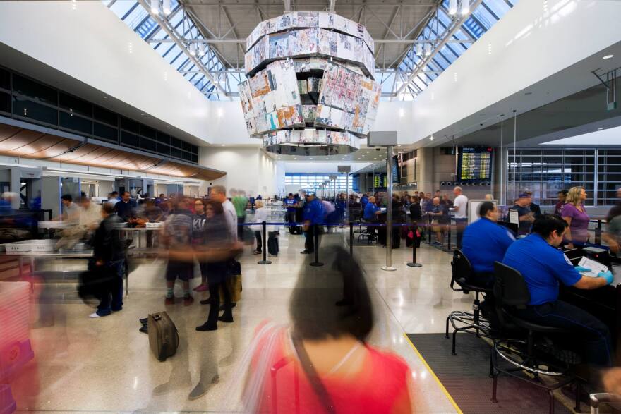A blurred view of the TSA room as dozens of people move through it. A large multicolor bell sculpture hangs in the air with panels of etched flat materials lined up to make the curve.