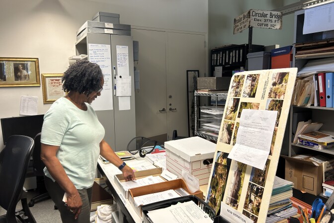 An older woman with dark brown skin, and short curly dark hair looks through papers on a table under fluorescent lighting in a small room. 