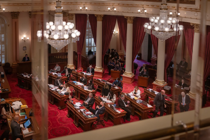 A legislative chamber with red carpeting, ornate columns, and crystal chandeliers with lighted globes. Nine long desks for two persons each are situated in rows of three each, with a 10th desk in a separate row at the rear. About a dozen legislators are seated at the tables, some reading on their phones, others reading paperwork scattered on the desks.
