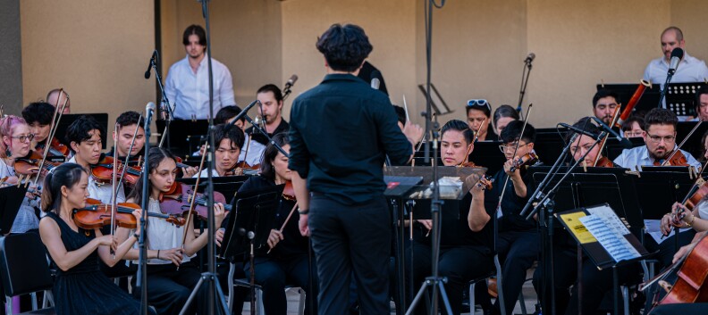 About 20 people sit in front of music stands. They hold musical instruments.