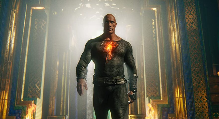 'The Rock' Johnson stands in the center of the image, mid-stride. He wears a skin-tight suit that is dark gray and covers his chest, arms, and down through his legs. The suit has a cracked texture, like lava rock or charred wood. A large, bulky leather belt sits around his waist, the same color of the suit, but a smoother texture. In the middle of Johnson's chest is a large orange lightening bolt that glows as if made of lava. Johnson stares straight ahead, eyes hooded. Behind him is an elaborate hallway with intricate wall designs of gold, emerald, and blue.