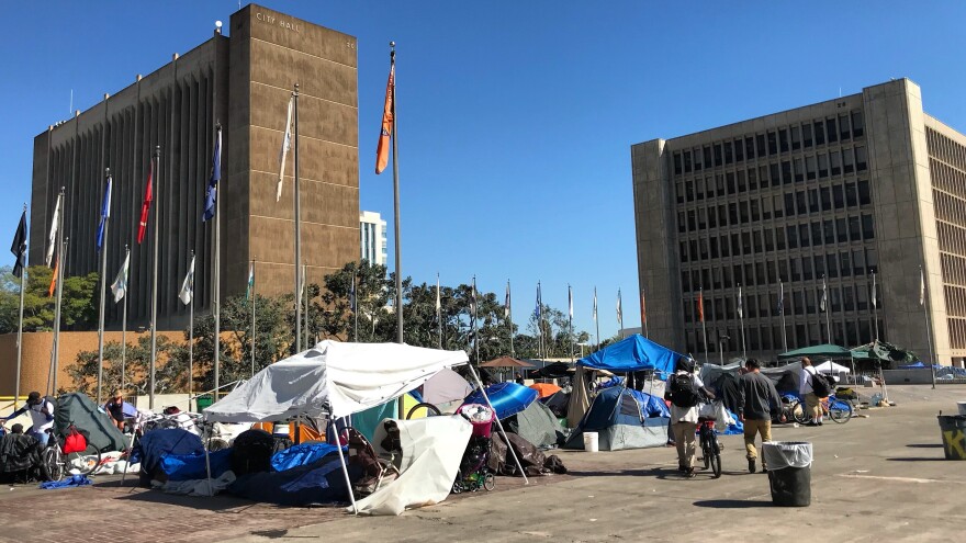 A homeless encampment sits in a lot in front of two multi-story buildings, and a clear, blue sky in the background. Flagpoles with different flags surround the lot. The flags are still.