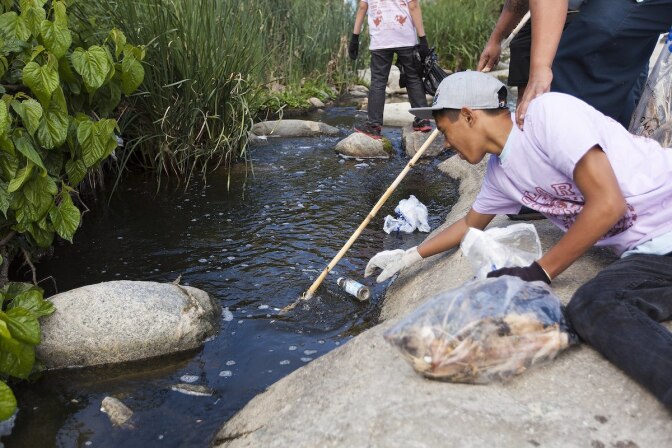 A volunteer in a pink shirt and gray hat reaches over to pick out a bottle from the LA River.