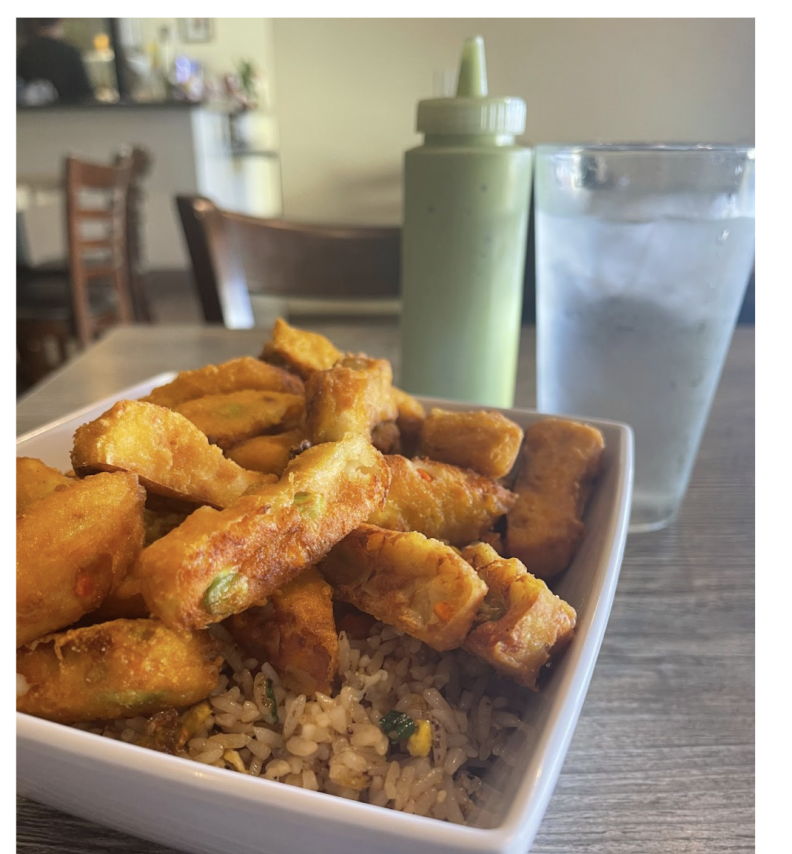 Rectangular fried cuts of tofu lay on top of a bed of brown fried rice in a square white bowl. Behind is a clear plastic squeeze bottle containing a light green sauce and a clear plastic cup of ice water