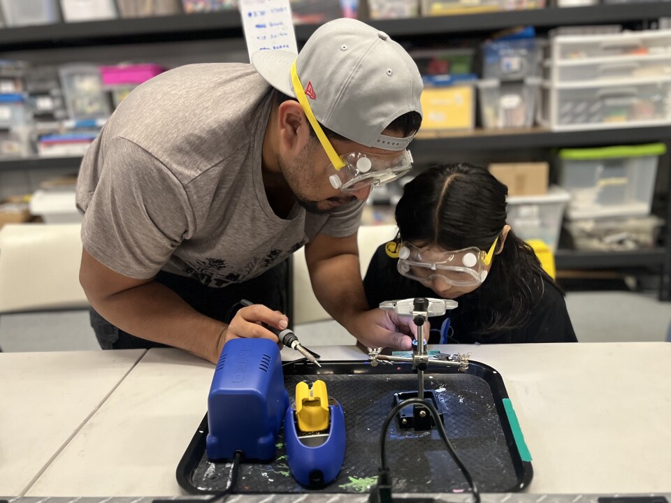 A man with medium skin tone helps his daughter use a soldering machine. Both are wearing safety glasses. He is wearing a baseball cap turned backward.