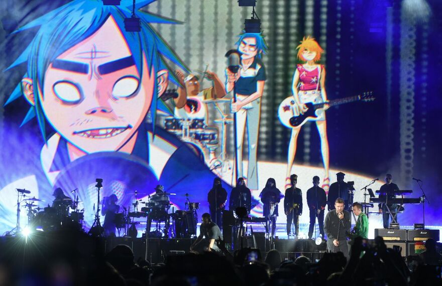 The animated images of the band Gorillaz appear on a background, showing a blue-haired figure at a mic, a female figure with orangeish hair on guitar, and a brown-skinned muscly man on drums. Below them on stage, the real life performers who make up this band perform, with other band members supporting them in the shadows behind.