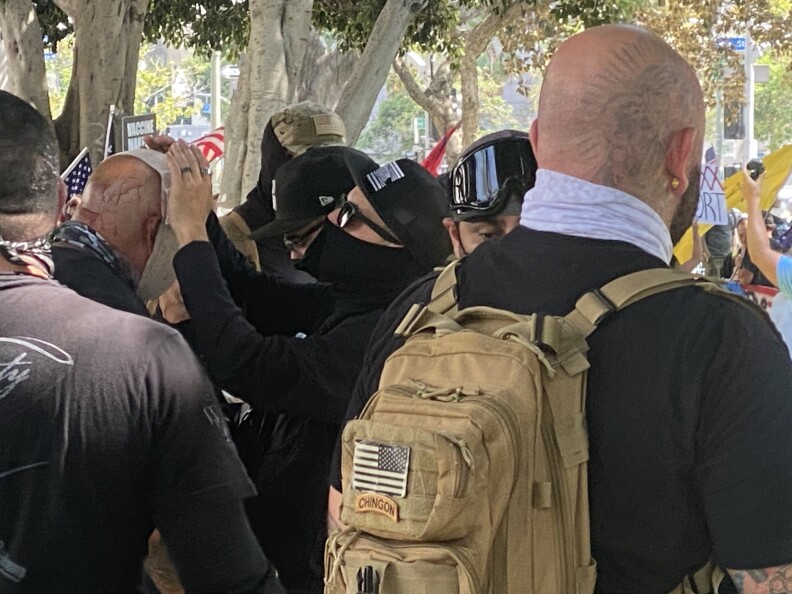 At an anti-vaccination rally in downtown LA on Aug. 14, 202, a group of men is clustered tightly together, with trees in the background. Two men in the foreground have their backs to the camera; the man on the right is wearing a small tan backpack with a small American flag on it. In the middle of the picture a man with a dark cap, sunglasses and a dark face mask is attending to a bald man with a bandage on his head. Blood is visible on the back of the man's head.