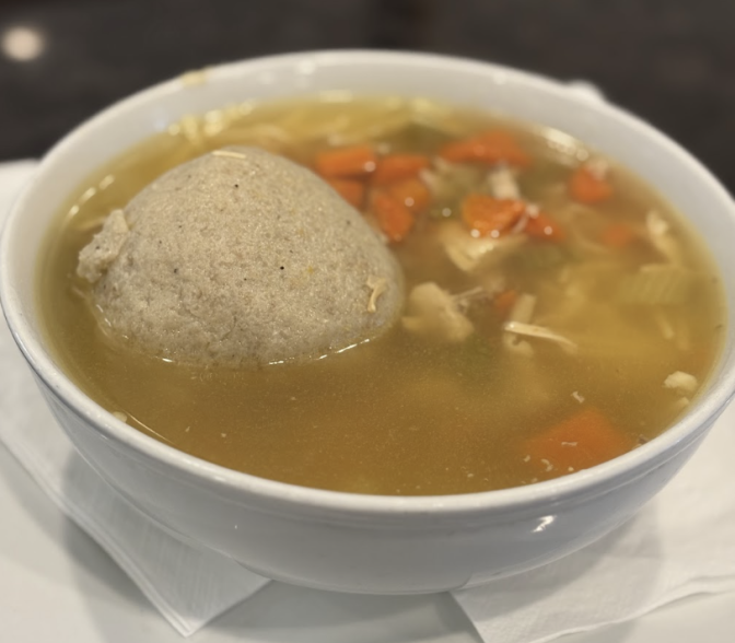 A bowl of matzo ball soup shows a deep delicious broth, a large matzo ball and assorted carrots and piece of chicken