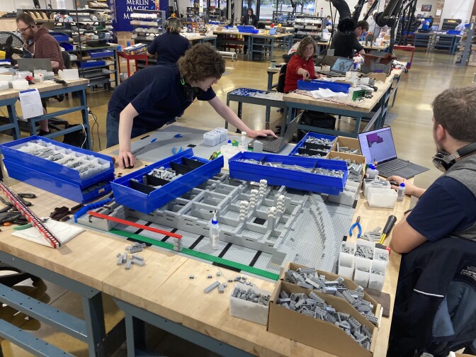 People at various work tables assemble Lego structures using Legos pulled out of various bins.