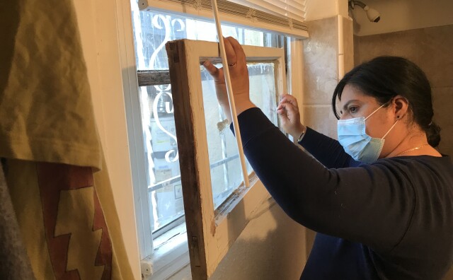 A woman wearing a mask in her apartment bathroom removes a broken window from its frame.