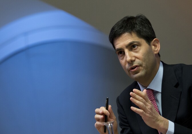 An angled view Kevin Warsh, a man with light skin tone, wearing a black suit and spotted tie, speaks while gesturing with both hands.