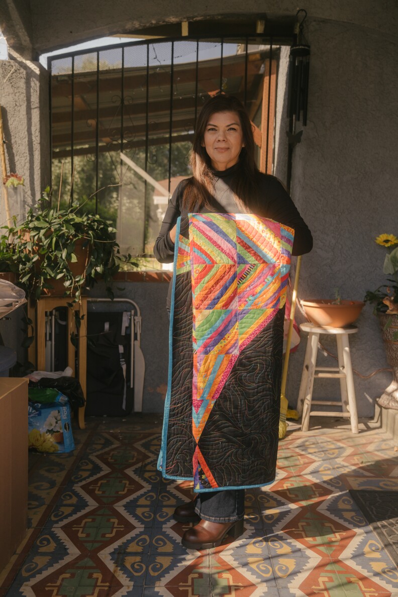 A woman with brown skin tone stands in daylight, carrying a colorful quilt hanging over her arms.