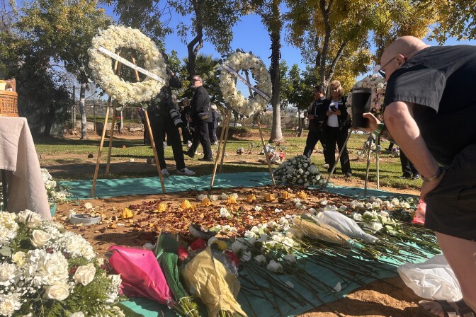 A man wearing all black t-shirt and shorts is leaning over a large communal grave buried in the dirt. He has a black coffee cup in his hand, outstretched towards the grave as if in a toast. The grave is covered in white roses and flower bouquets. 